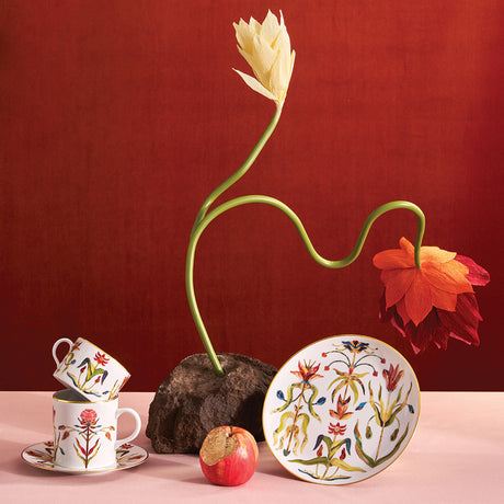 Decorative floral arrangement with a twisted stem, teacup, saucer, and plate on a red background.