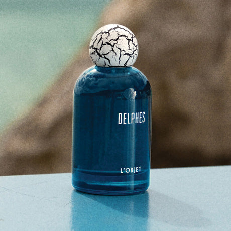 Blue perfume bottle with cracked cap labeled 'Delphes' by 'L'Objet' on a blurred background