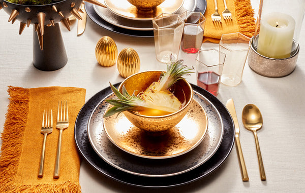 Radiant table setting with black, platinum and gold glazed Alchimie plates, gold creal bowl with cut pineapple, gold hexagonal glasses, gold Carrousel salt and pepper shakers.