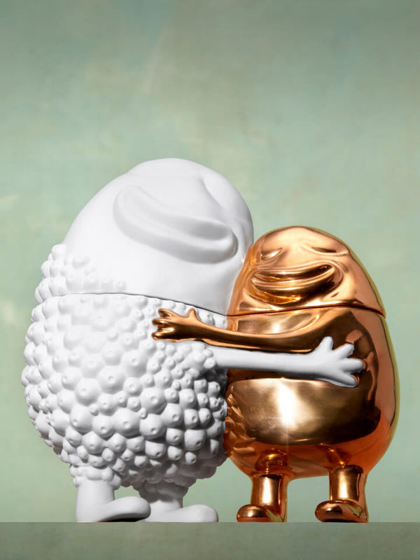 Two egg-shaped decorative sculptures standing side by side, one matte white with a bumpy textured surface and one metallic gold with a smooth reflective finish, posed against a soft pale green background.