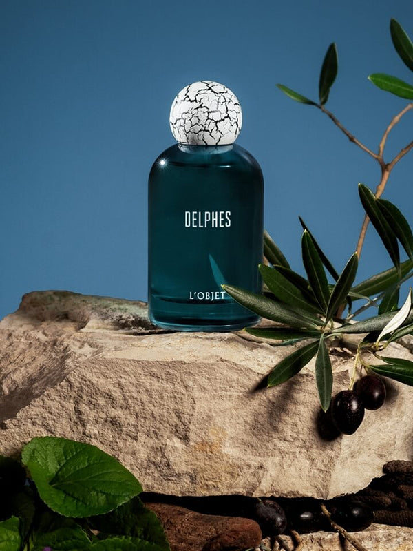 Blue perfume bottle labeled 'Delphes' on a stone surface with olive branches and olives.