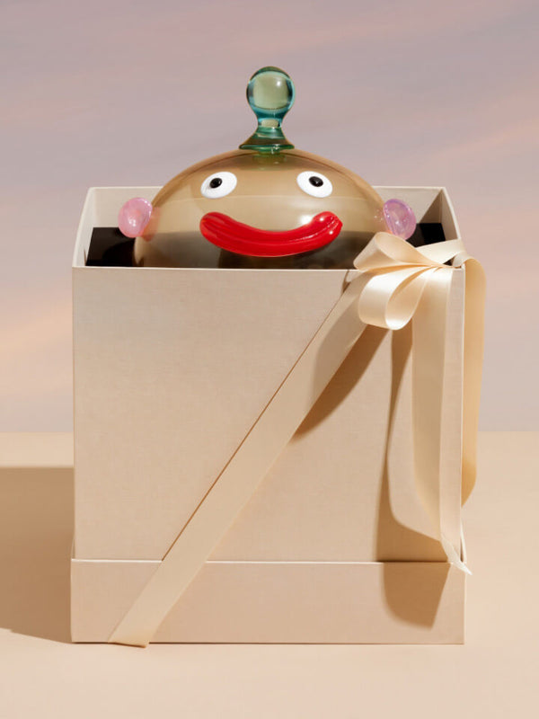 A ceramic character figurine with a red smile, pink ears, and a green top knob, presented inside a cream-colored gift box with a ribbon.