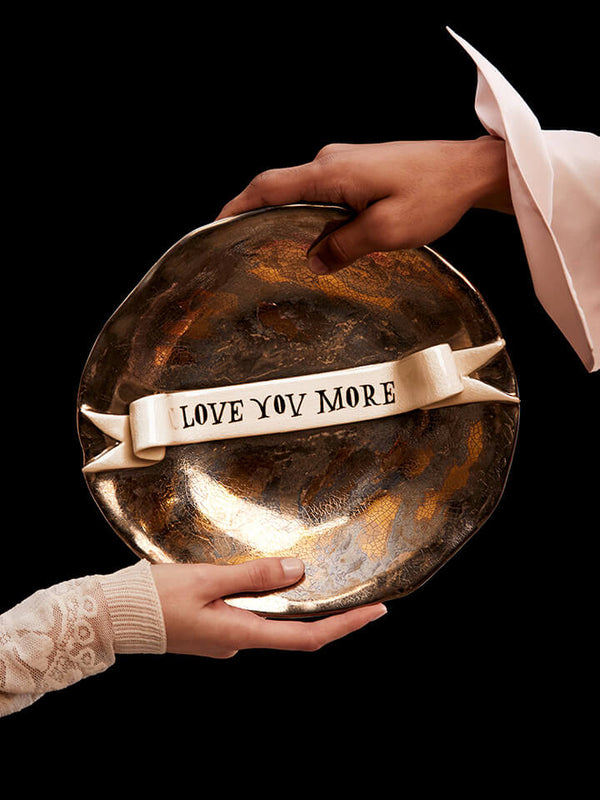 Metallic tray with 'LOVE YOU MORE' text held by two hands against a black background